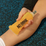 A hand holding a filled NOBONK Pineapple Mango soft flask against a leg on a blue athletics track