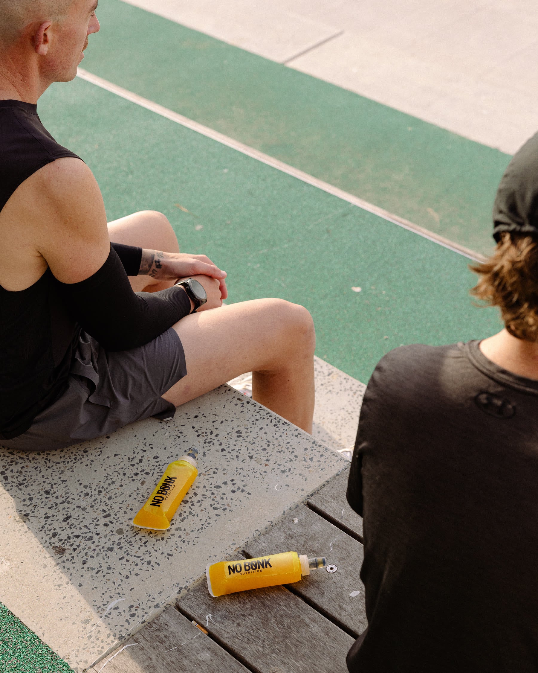 Two runners sitting on a boardwalk after a session, filled NOBONK flasks on the ground beside them